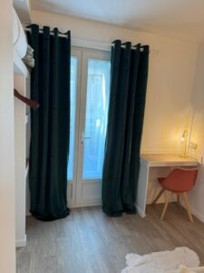 Chambre moderne avec lit mezzanine, bureau en bois clair, chaise design orange, lampe dorée et rideaux vert foncé. Sol stratifié clair.