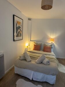 Chambre cosy avec lit double, parure beige, oreillers corail. Quatre serviettes roulées, lampes de chevet, tableau tropical et parquet.