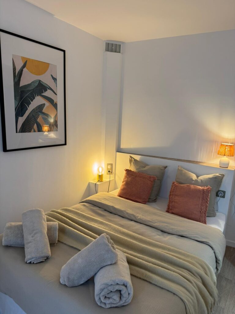 Chambre moderne cosy : lit double, draps beiges, coussins terracotta, serviettes. Lumières chaudes, tableau tropical encadré.