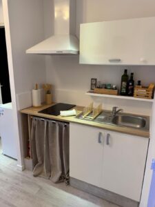 Cuisine moderne blanche et bois clair avec hotte, plaque induction, évier, ustensiles. Un rideau gris cache un rangement sous le plan de travail.