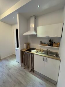 Cuisine moderne équipée d'un appartement ou studio: plan de travail bois clair, évier, plaque de cuisson, hotte, placards blancs et rangements rideau. Réfrigérateur visible.