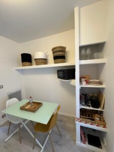 Cuisine/salle à manger moderne avec table verte, chaises, étagères murales, micro-ondes, vaisselle et paniers tressés.