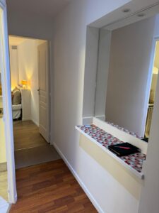 Couloir lumineux avec miroir mural au-dessus d'une étagère carrelée (rouge, gris, blanc), un carnet et stylo. Porte ouverte sur une pièce éclairée par une lampe chaude. Sol en parquet.