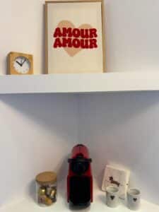Étagères blanches avec machine Nespresso rouge, capsules, deux mugs cœur, et serviettes "Je t'aime". Dessus, horloge bois et cadre "AMOUR AMOUR" cœur rose.
