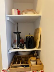 Étagères de cuisine blanches avec bouilloire, grille-pain noir (bouton CANCEL), vaisselle empilée, mug, couverts en bois et sucre sur carrelage mosaïque.