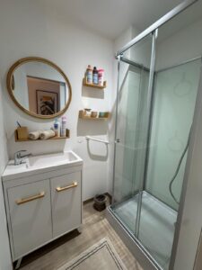 Salle de bain moderne blanche avec meuble vasque, miroir rond en rotin, étagères en bois, produits Mixa, Aria, Novog, Cadum, et douche vitrée.