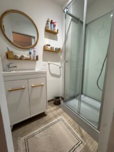 Salle de bain moderne et lumineuse. Miroir ovale, meuble vasque blanc et bois, étagères murales avec produits d'hygiène Mixa, Cadum. Douche vitrée et tapis beige.
