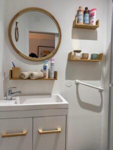 Salle de bain moderne: lavabo, miroir rond rotin, étagères bois. Produits Mixa, ARTA, Franck Provost, Cadum. Barre d'appui murale.