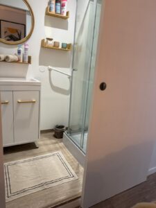 Salle de bain moderne: meuble vasque blanc, miroir rond, douche vitrée, étagères bois. Produits Mixa, Cadum, Provost. Tapis de bain beige.